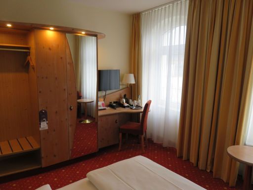 Mercure Hotel Berlin Mitte, Deutschland, Zimmer 306 mit Kofferablage, Schrank mit Minisafe, Spiegel, Flachbildfernseher, Tisch, Stuhl, Kühlschrank und Wasserkocher